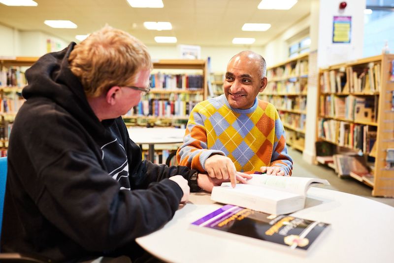 Two adult students sat at a table discussing a book and smiling