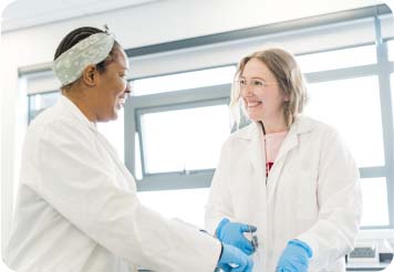 Two students in science lab wearing white lab coats and gloves looking at each other and smiling