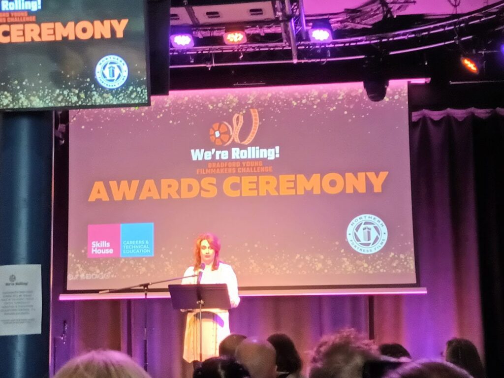 A lady in a white dress stands in front of a podium on a stage. Behind her is a screen that says 'We're Rolling Bradford Young Filmmakers Challenge Awards Ceremony'.