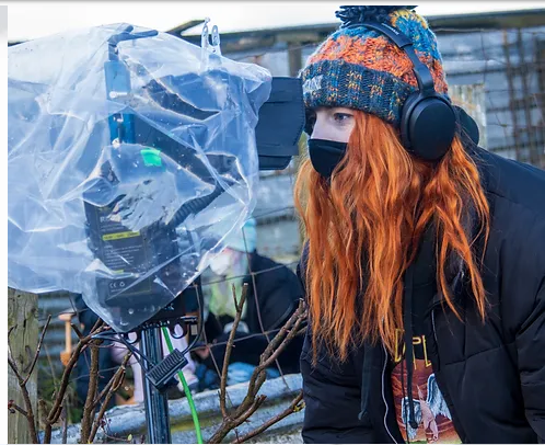 A woman with long, red hair looks into a video camera on the set of a film