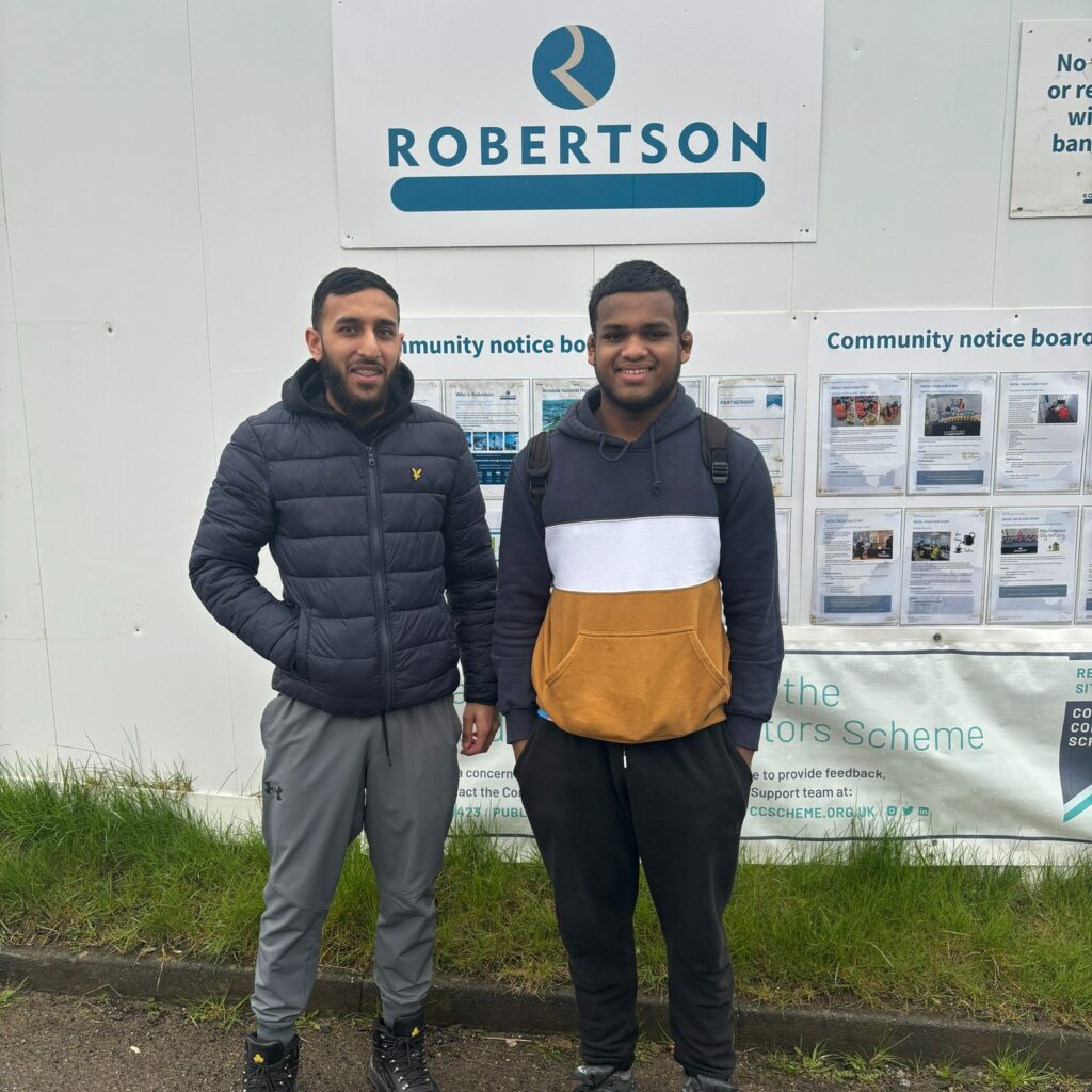 Two young men smile for the camera whilst standing in front of a sign that says, 'Robertson'