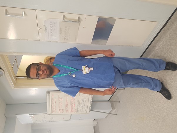 A man wearing glasses, a lanyard and hospital scrubs stands on a hospital ward.