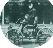 Black and white image of a man sat on a horseless carriage