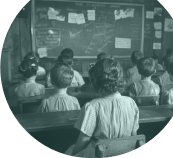 Black and white image of an old fashioned classroom with students sat facing a blackboard