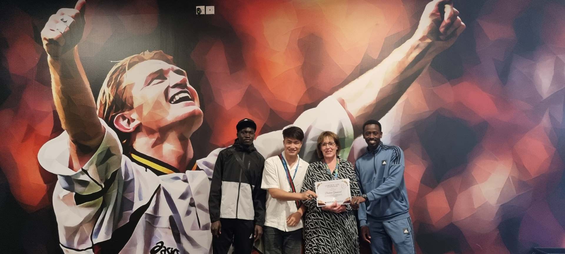 Three students and a member of staff posing with the award in front of a football mural