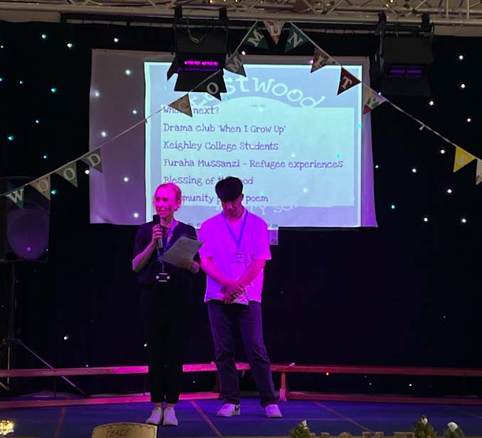 A Keighley College student on stage with host at Eastwood Primary School talking about their journey to the UK