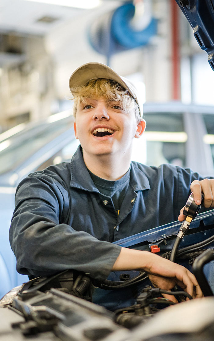 Motor vehicle student wearing a cap and smiling whilst working under a car bonnet