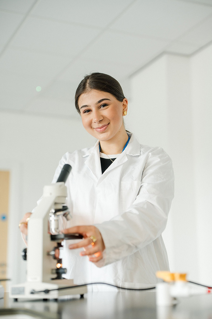 Student in lab coat smiling whilst using a microscope in the Science lab